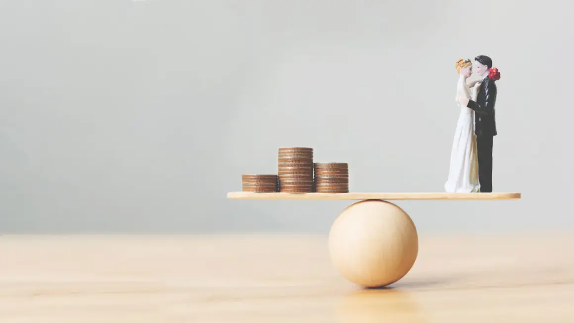 Figurines de mariés en équilibre sur une balance avec des pièces. Une métaphore visuelle pour comment préparer un mariage et gérer son budget.