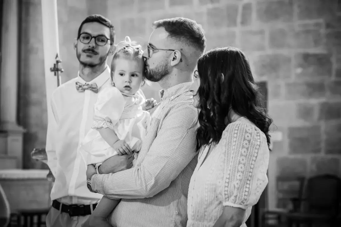 Un père embrasse sa fille, une petite fille en robe blanche, lors de son baptême. La question "jusqu'à quel âge peut-on baptiser ?" se pose.
