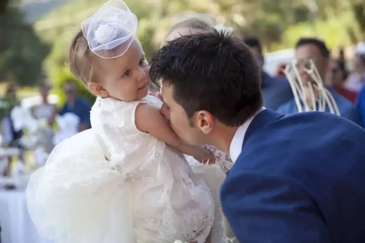 Un homme embrasse la joue d'un bébé en robe blanche, peut-être lors d'un mariage et baptême le même jour.