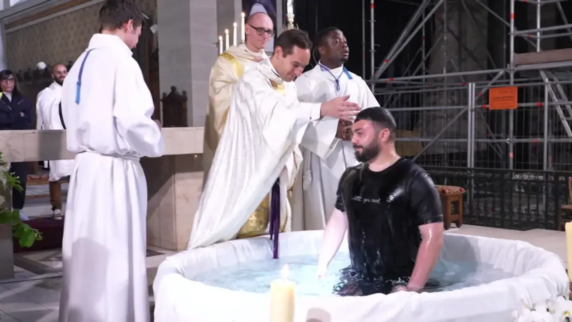 Cérémonie de baptême adulte dans une église. Un homme en t-shirt noir est immergé dans l'eau d'une cuve, entouré de religieux en soutanes blanches.