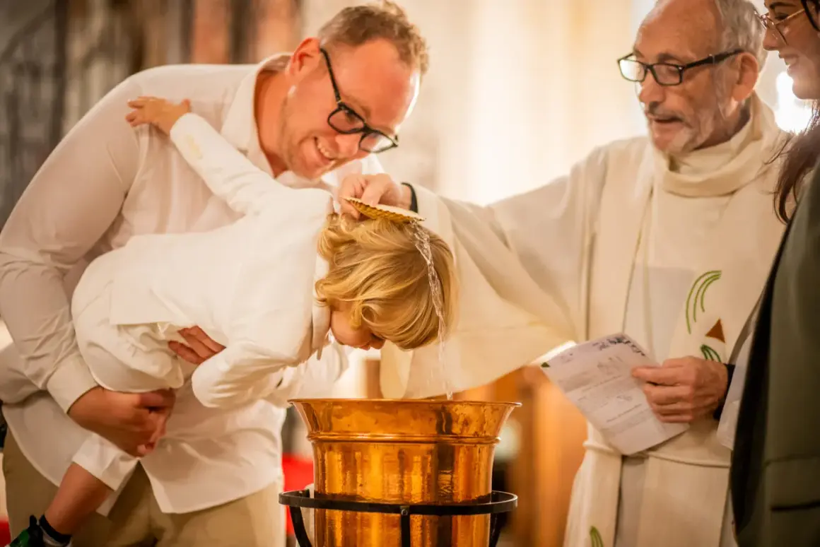 Cérémonie de baptême : un prêtre verse de l'eau sur la tête d'un enfant tenu par son père.