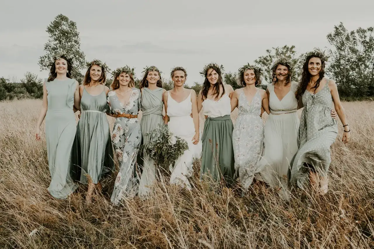 Un groupe de femmes souriantes, couronnées de verdure, célèbre un mariage champêtre. Elles portent des robes légères aux tons pastel, marchant dans l'herbe haute.