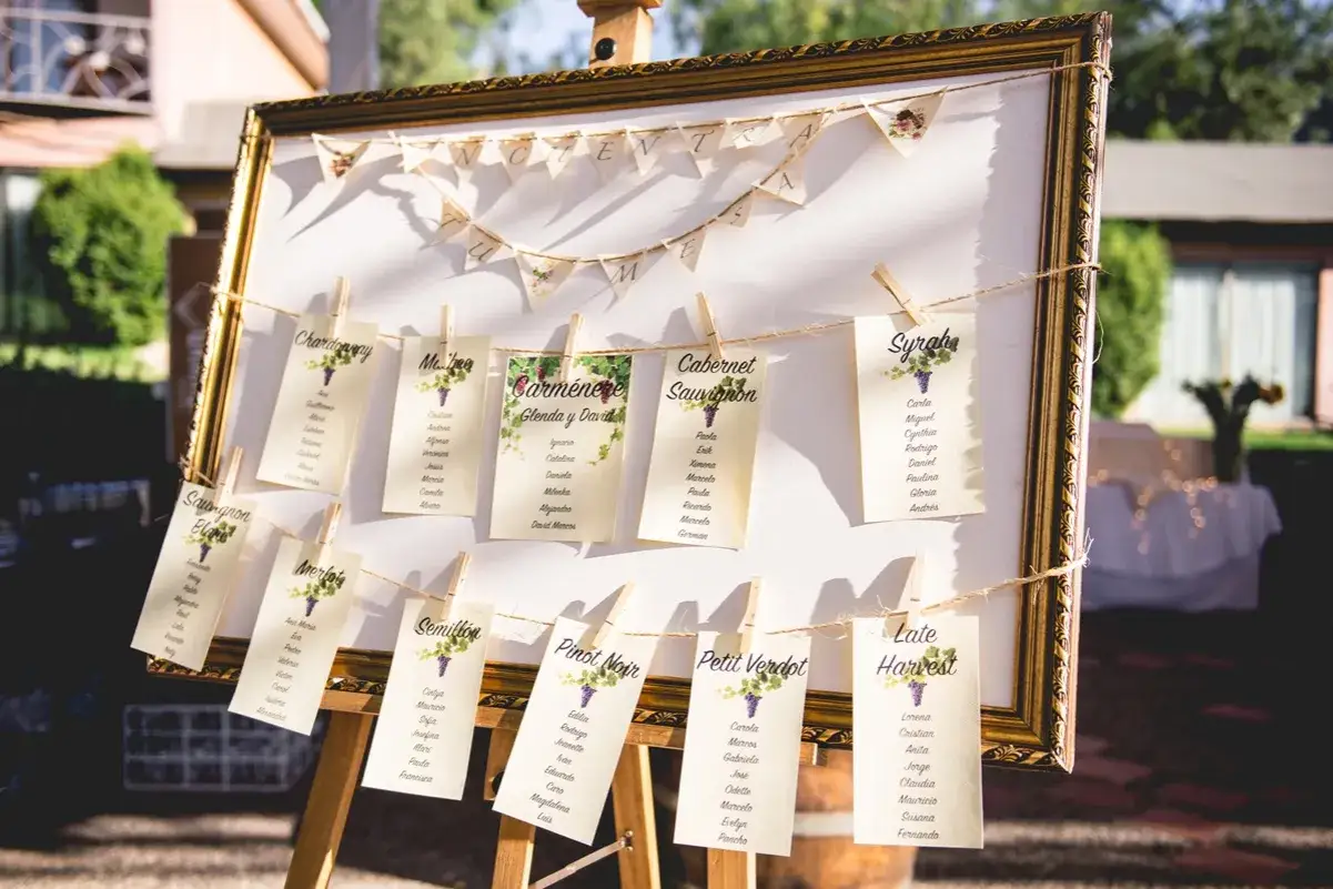 Un panneau doré présente un nom de table mariage avec des cartes suspendues par des pinces à linge, listant des noms d'invités sous des noms de vins.
