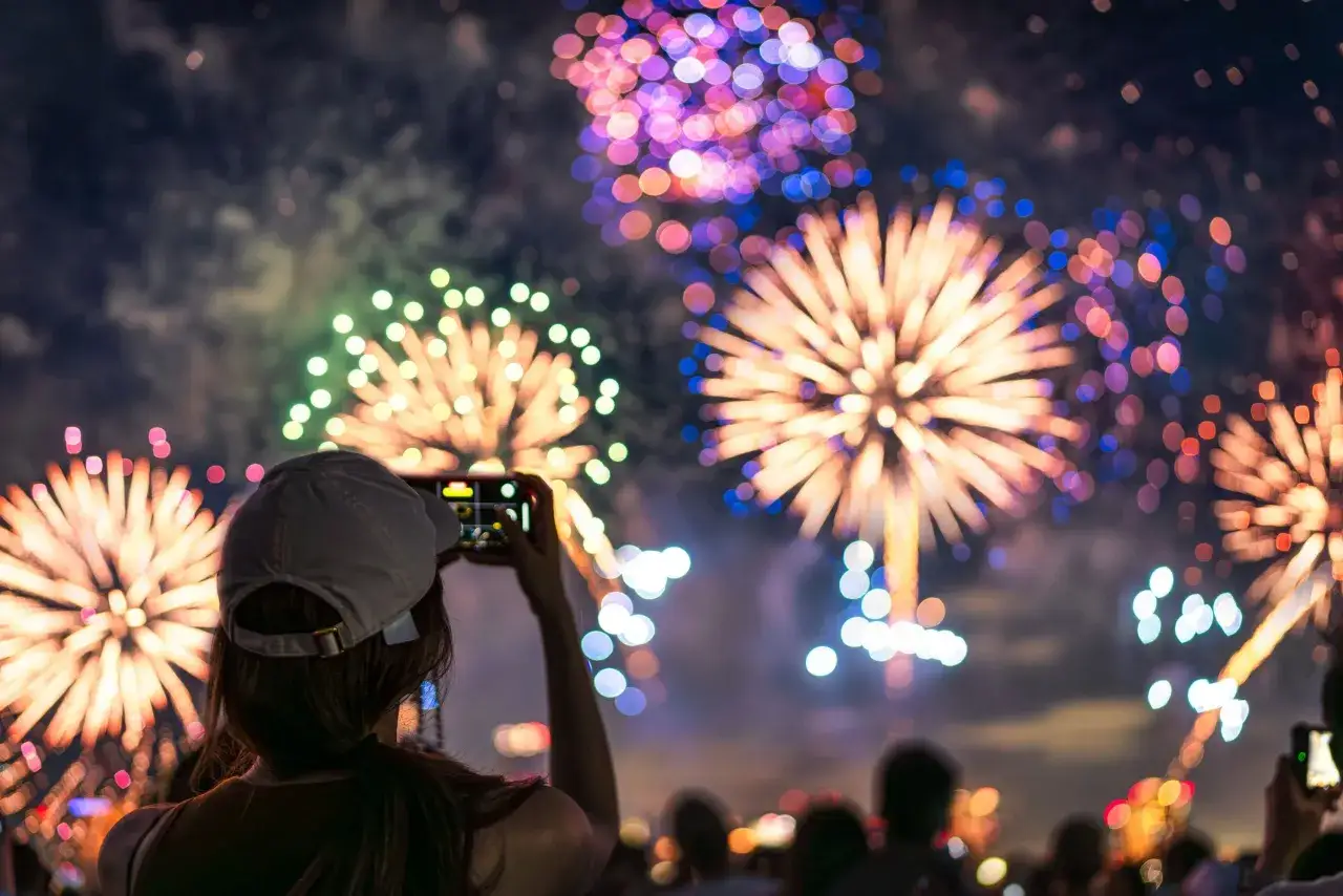 Une femme filme un magnifique feu d'artifice avec son t&eacute;l&eacute;phone, capturant la magie des explosions color&eacute;es dans le ciel nocturne.