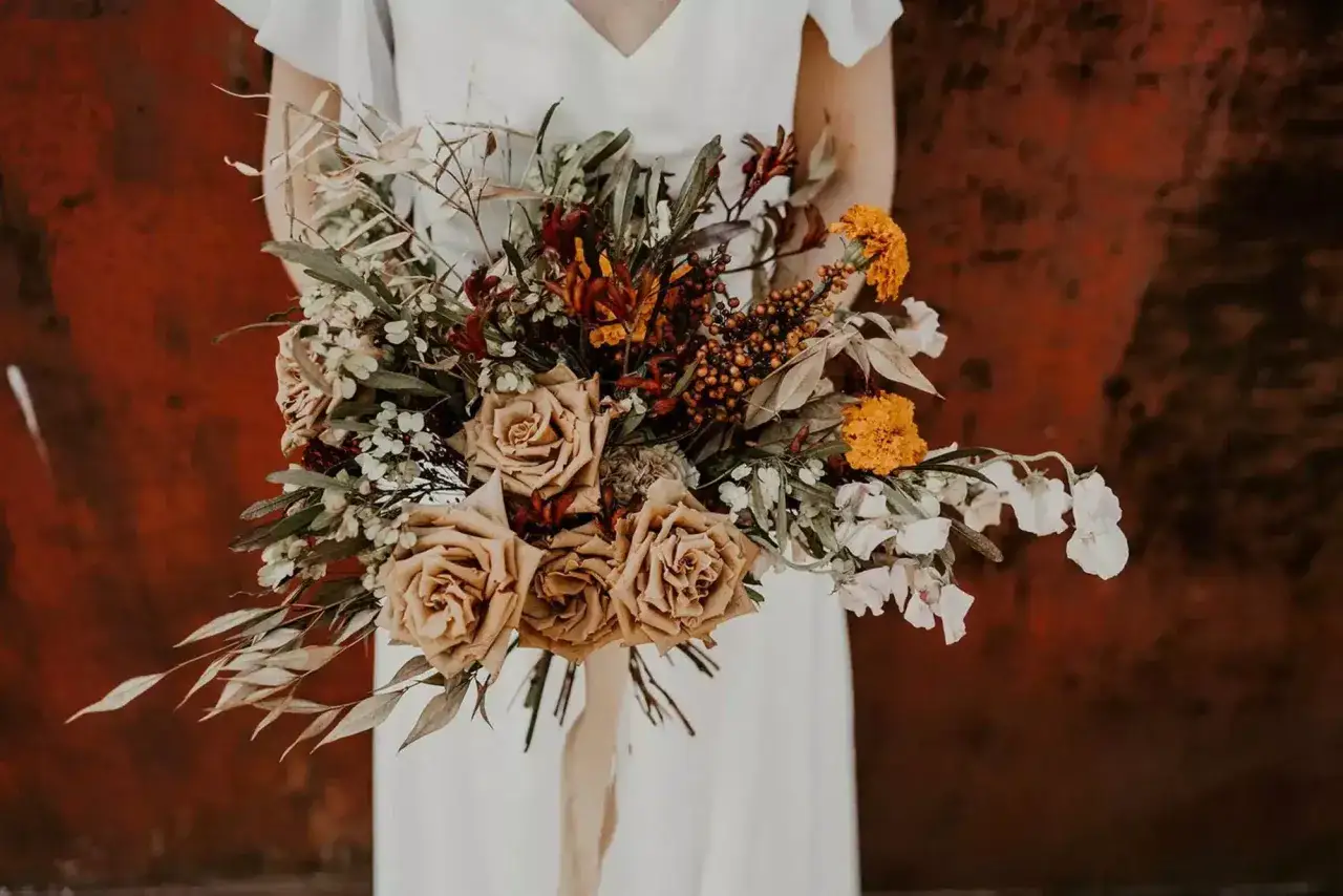 Un bouquet de mariée aux tons chauds, avec des roses saumon, des fleurs oranges et des feuillages secs. Une belle inspiration pour une liste des fleurs pour mariage champêtre.