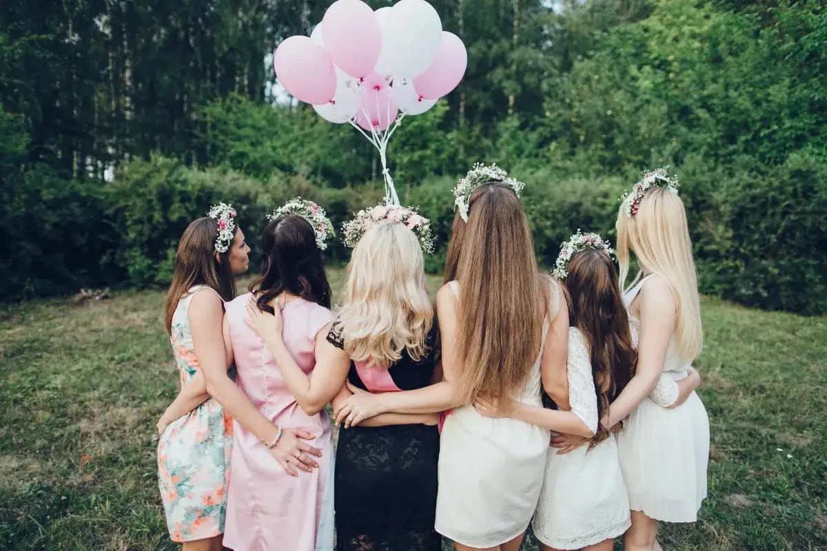 Amies en cercle, couronnes de fleurs, ballons roses et blancs. Un joyeux jeu EVJF en pleine nature.