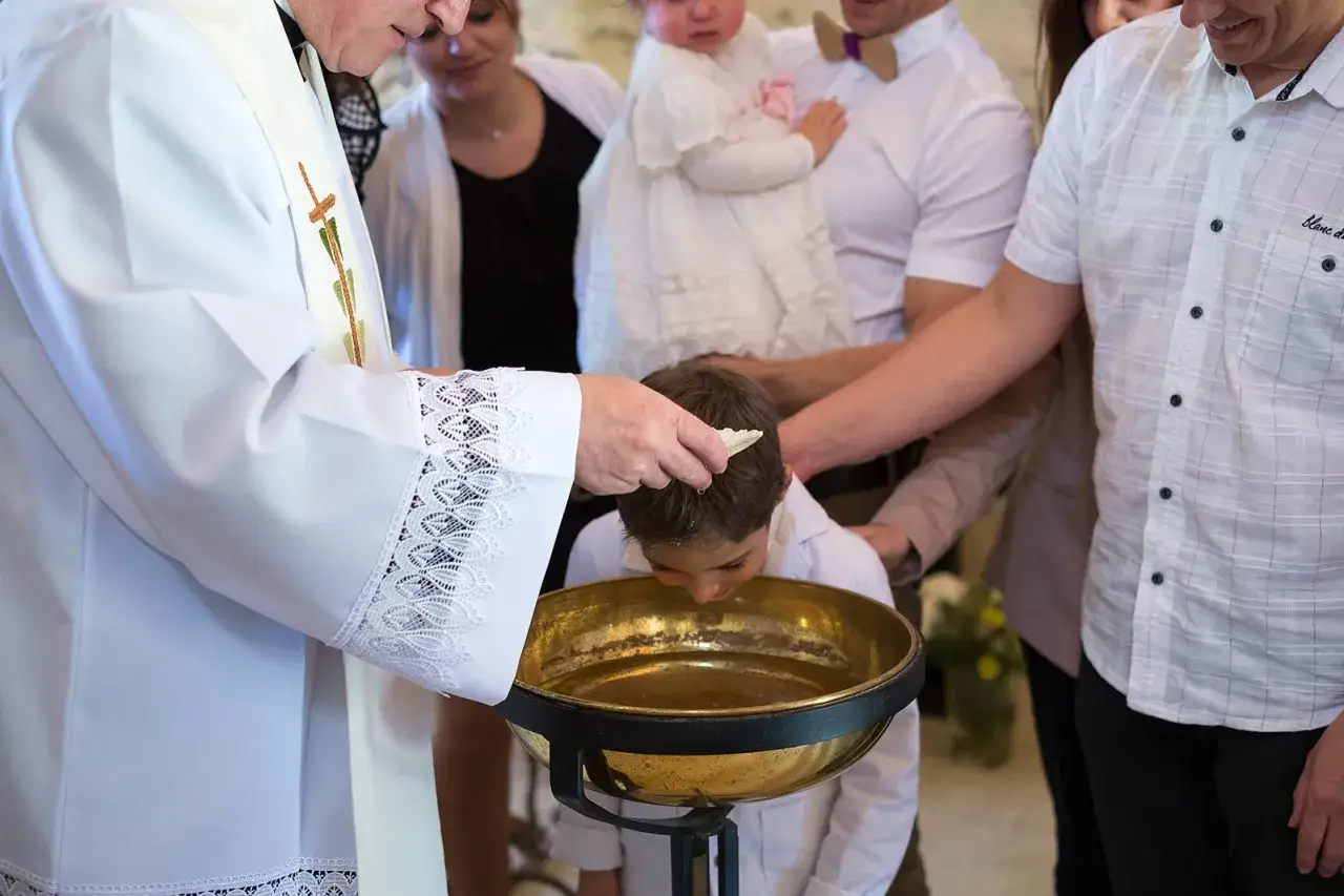 Un garçon de 10 ans se fait baptiser, penché au-dessus d'une vasque dorée, tandis qu'un prêtre verse de l'eau sur sa tête.