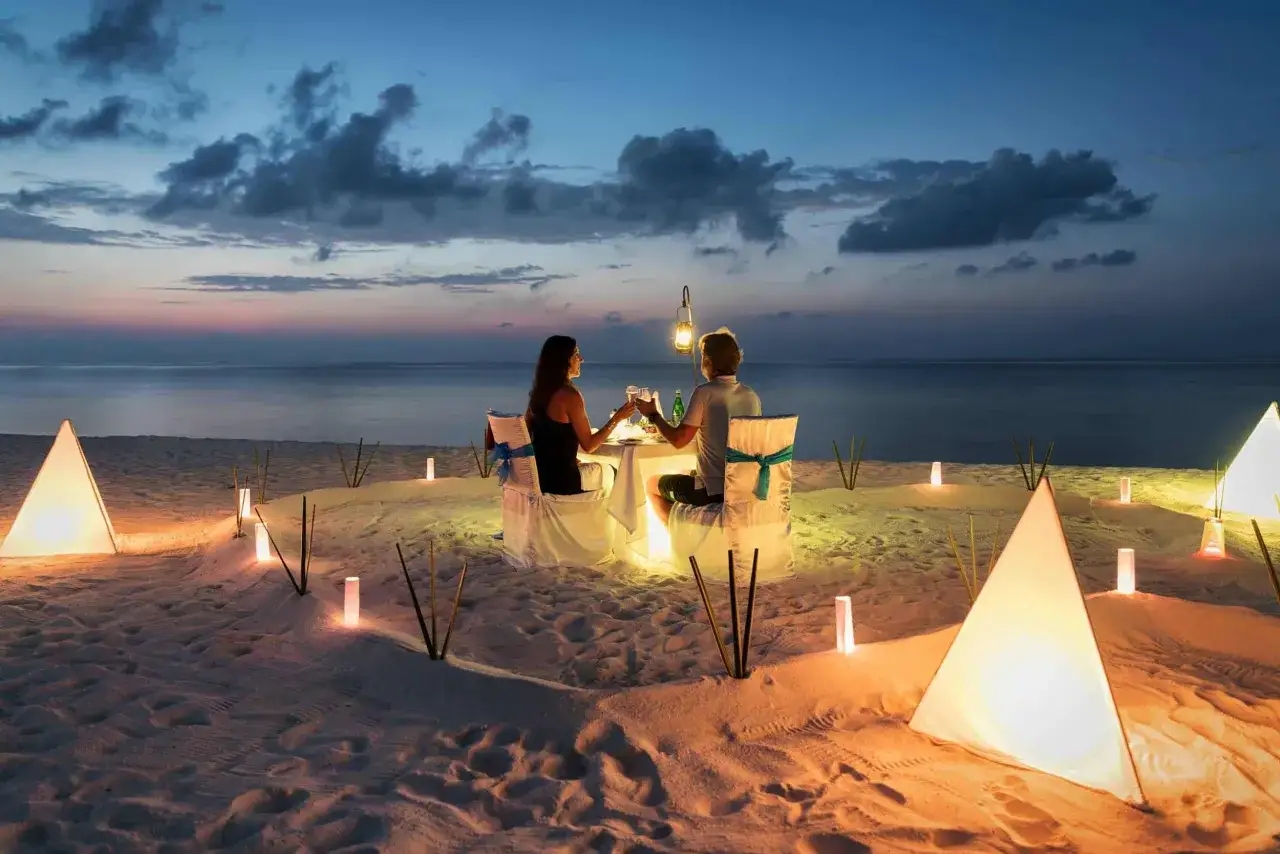 Un couple trinque lors d'un dîner romantique sur la plage, éclairé par des lanternes. Une parfaite destination lune de miel pas cher.
