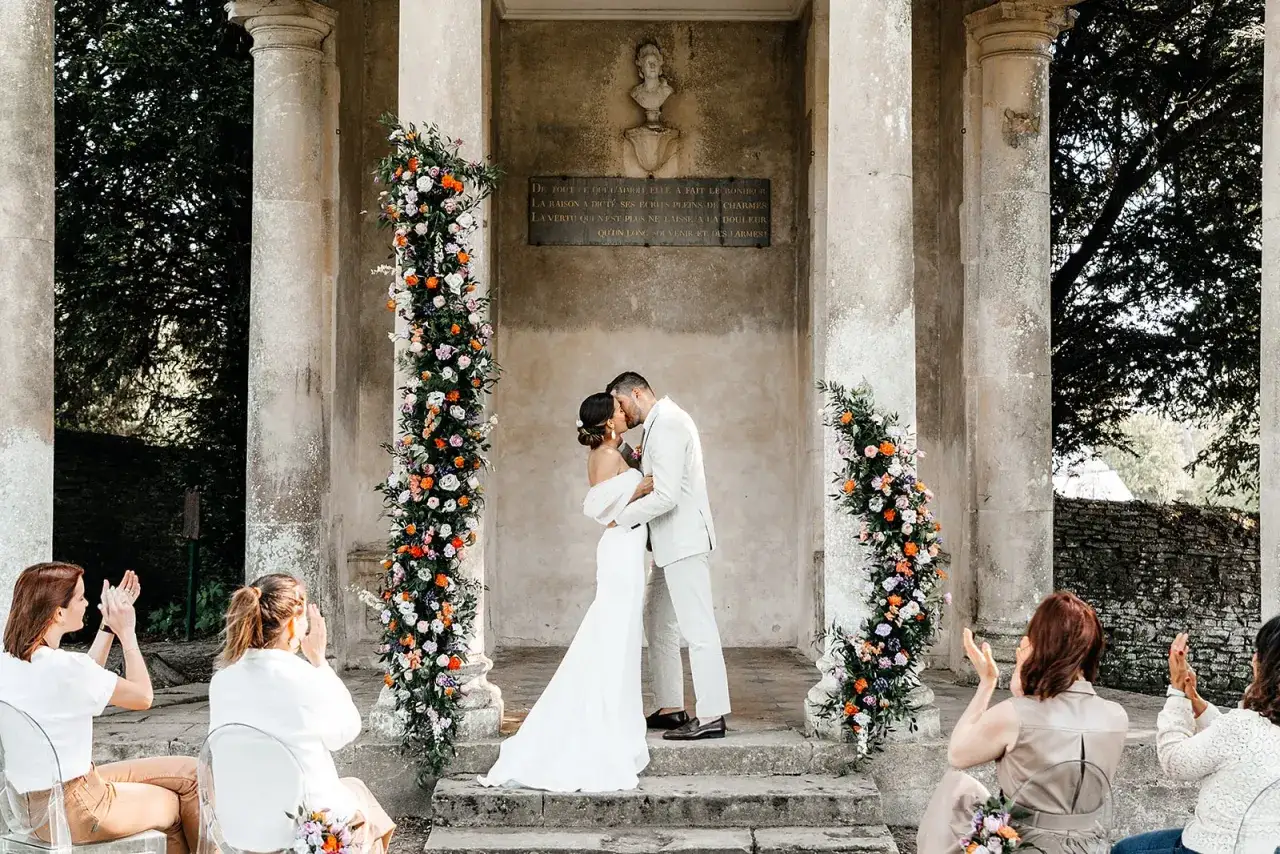 Un couple s'embrasse lors d'un mariage Bridgerton, entouré de fleurs colorées. Des invités applaudissent, créant une scène romantique et élégante.