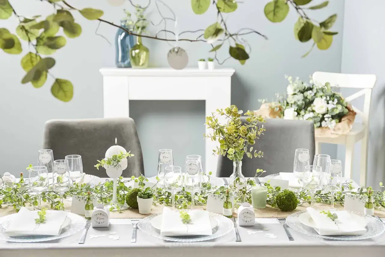 Une table joliment dressée pour organiser un baptême, avec des guirlandes vertes, des bougies et des verres gravés.