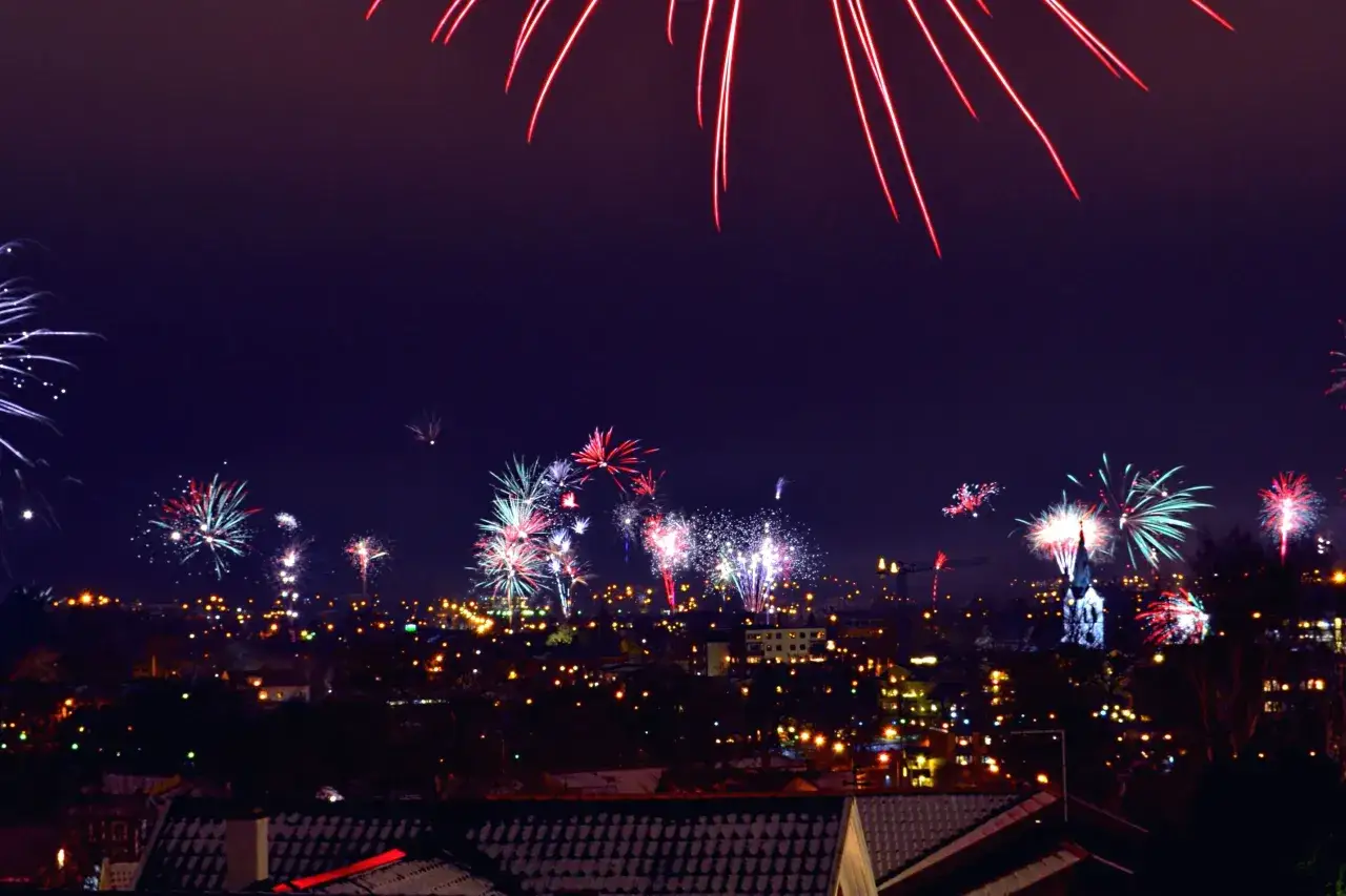 Un ciel nocturne illuminé par un feu d'artifice spectaculaire, des gerbes de lumière rouge et blanche éclatent au-dessus d'une ville endormie.
