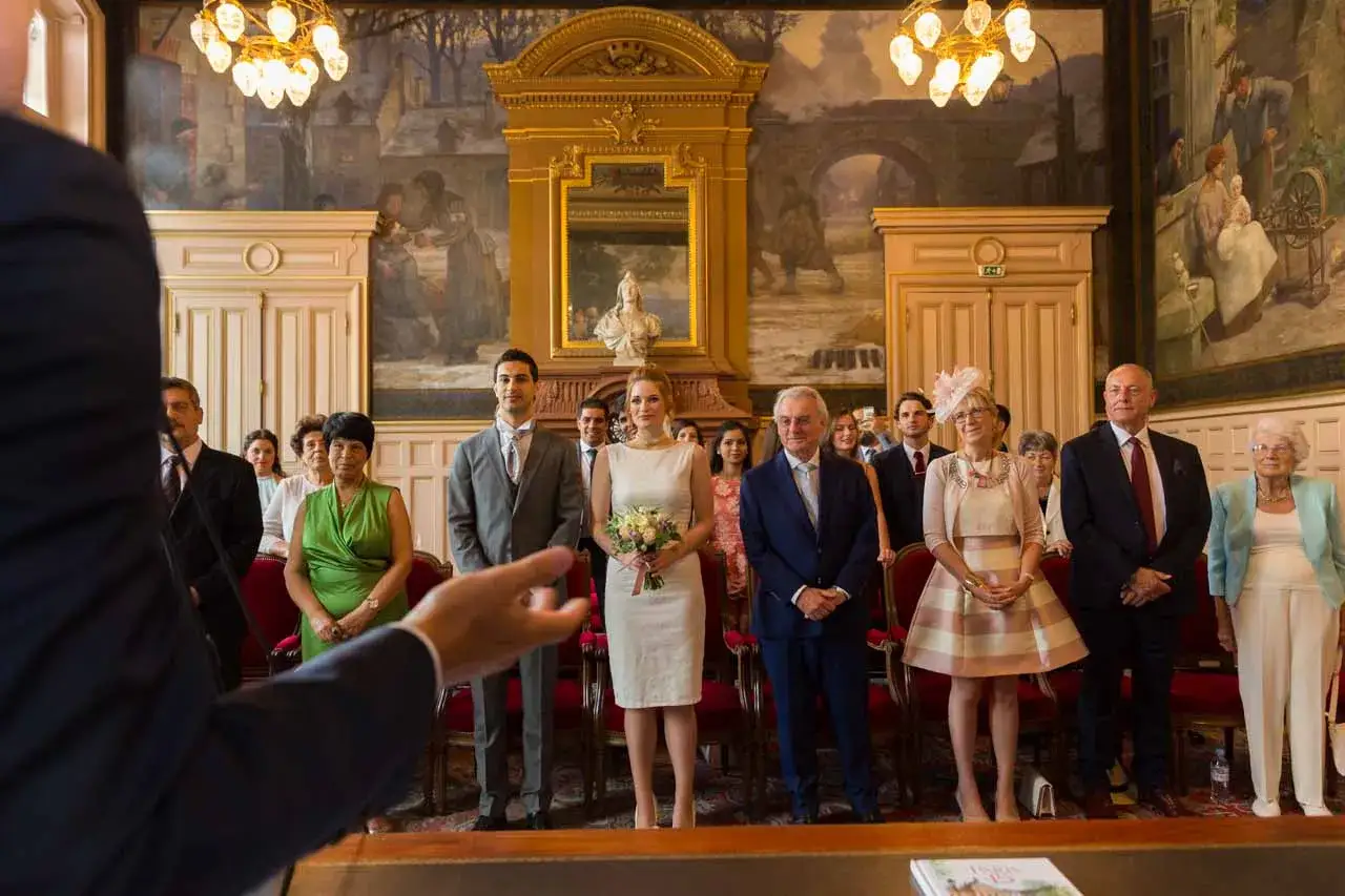 Cérémonie de mariage : le marié, la mariée avec bouquet, et plusieurs invités. On peut compter au moins 5 témoins.
