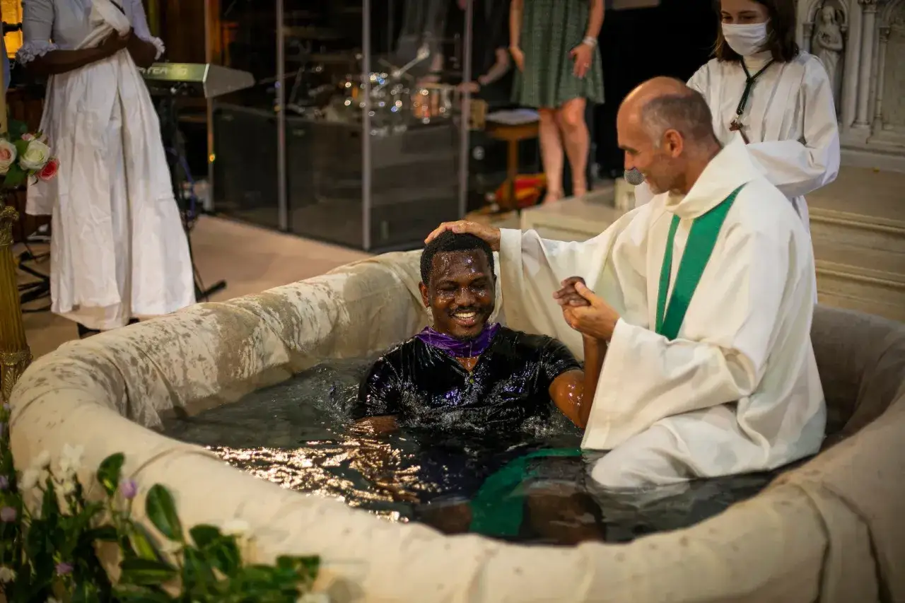 Un homme souriant est immergé dans l'eau pour un baptême sous condition, béni par un prêtre.