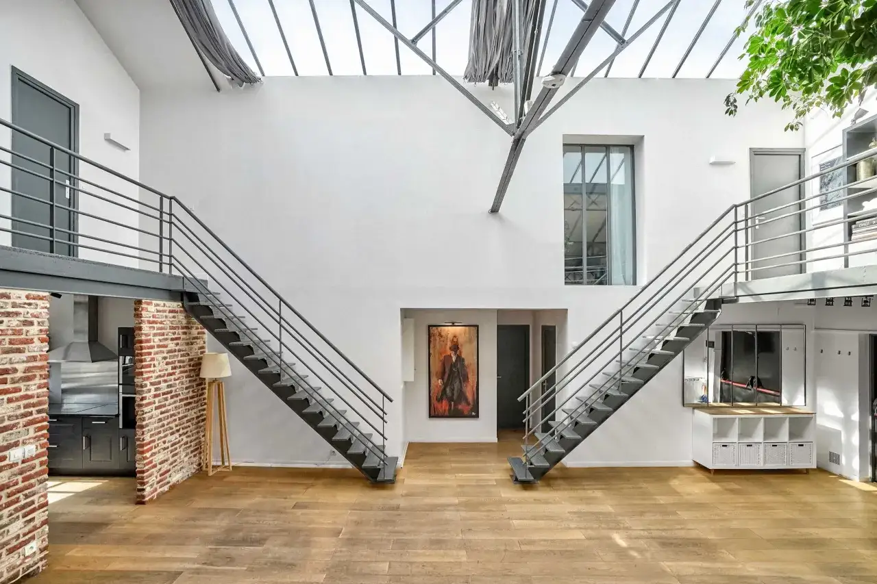 Escalier métallique menant à une mezzanine dans un loft au style mariage industriel. Le sol en bois clair contraste avec les murs blancs et la brique apparente.