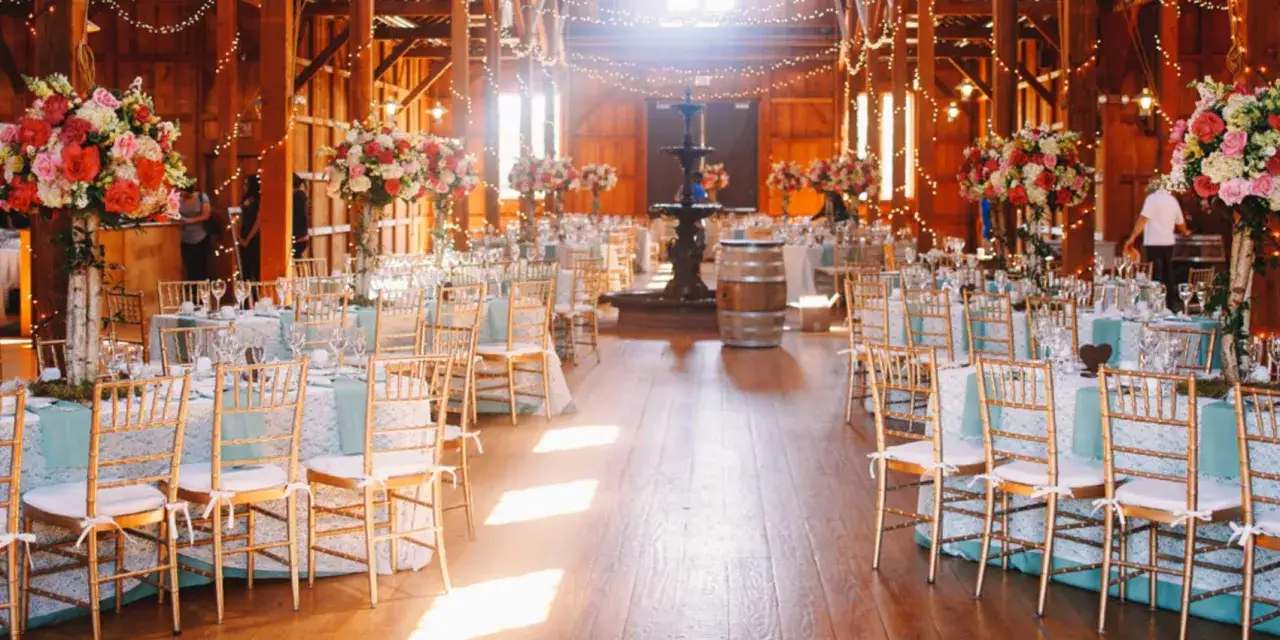 Disposition table mariage dans une grange rustique, décorée de fleurs roses et blanches, de guirlandes lumineuses et d'une fontaine.