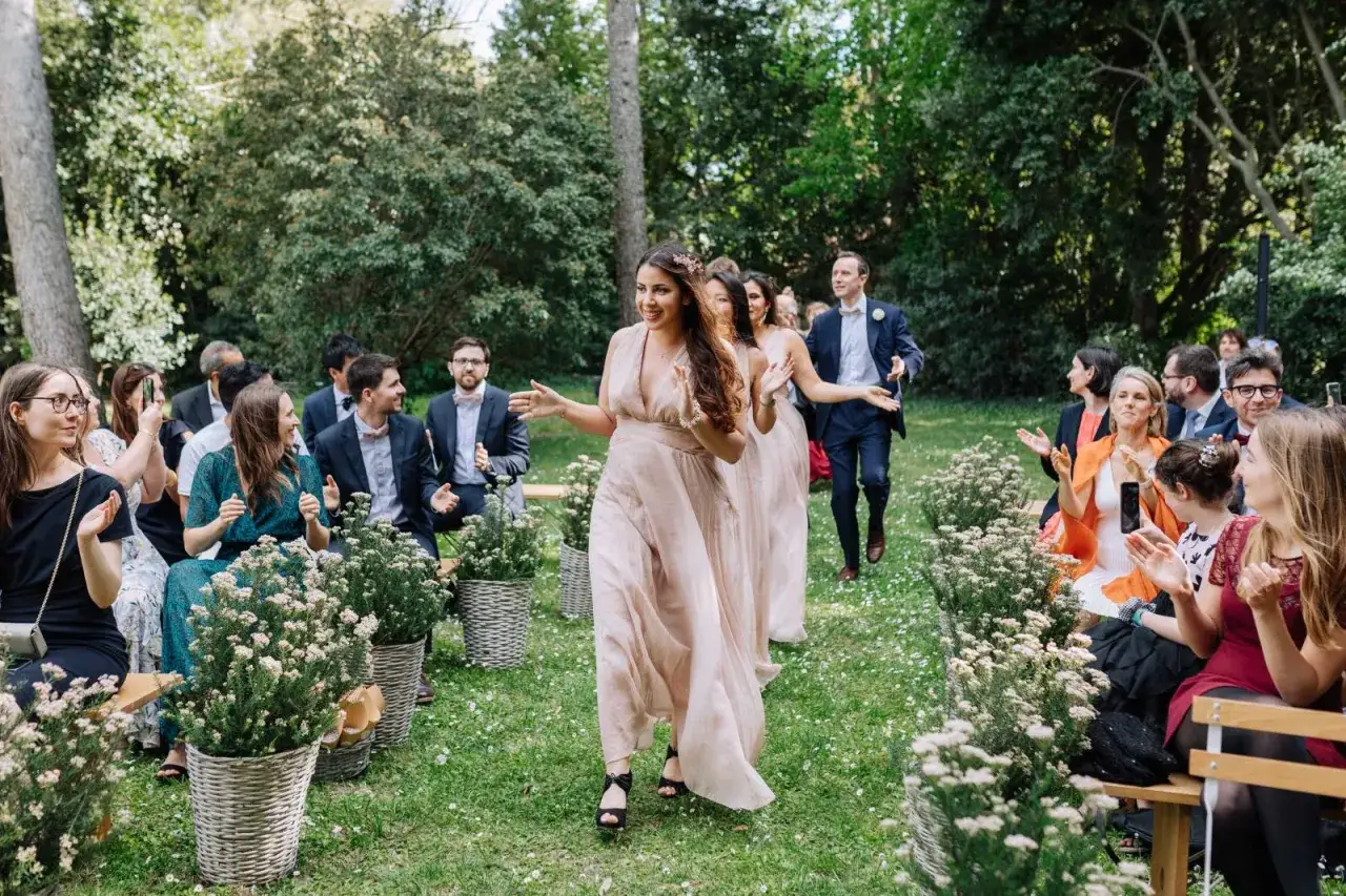 Le cortège mariage avance joyeusement dans un jardin fleuri. Les invités applaudissent, ravis.