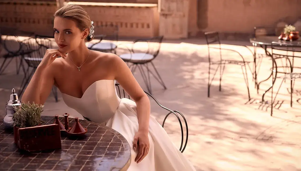 Une mariée en robe bustier, parée d'un collier délicat, attend à une table extérieure. Son style épuré évoque un mariage nude, empreint d'élégance naturelle.