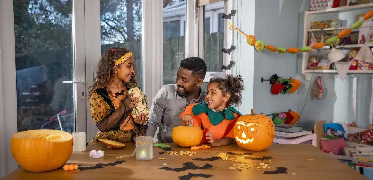 Un père et ses deux filles préparent leur thème soirée halloween. Citrouilles sculptées, costumes et décorations créent une ambiance festive.