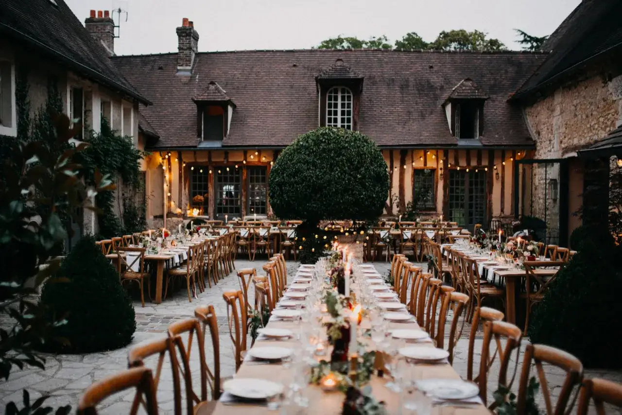 Tables dressées pour un mariage campagne, éclairées par des guirlandes lumineuses dans la cour d'une bâtisse ancienne.