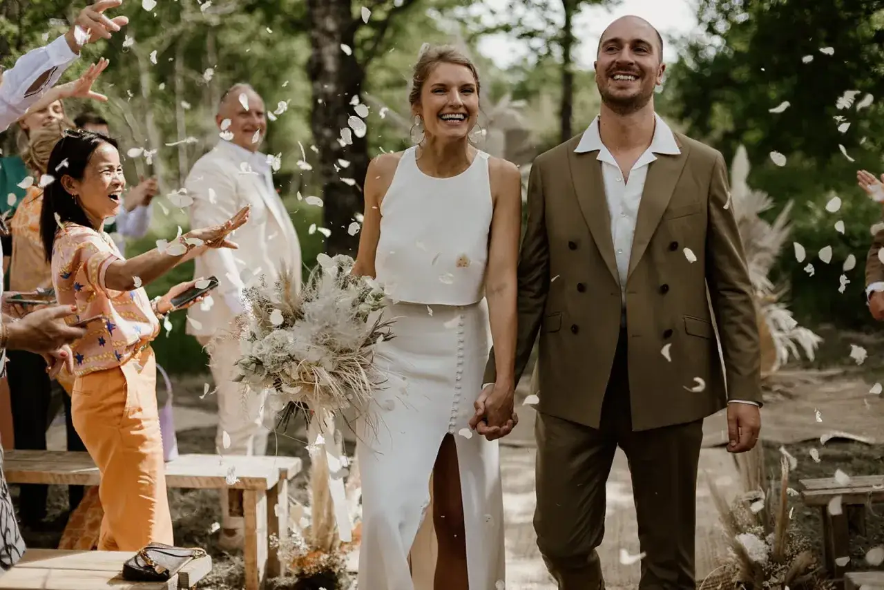 Un couple rayonnant traverse une pluie de pétales lors d'une cérémonie laïque originale. Les invités lancent des confettis, créant une atmosphère joyeuse et festive.