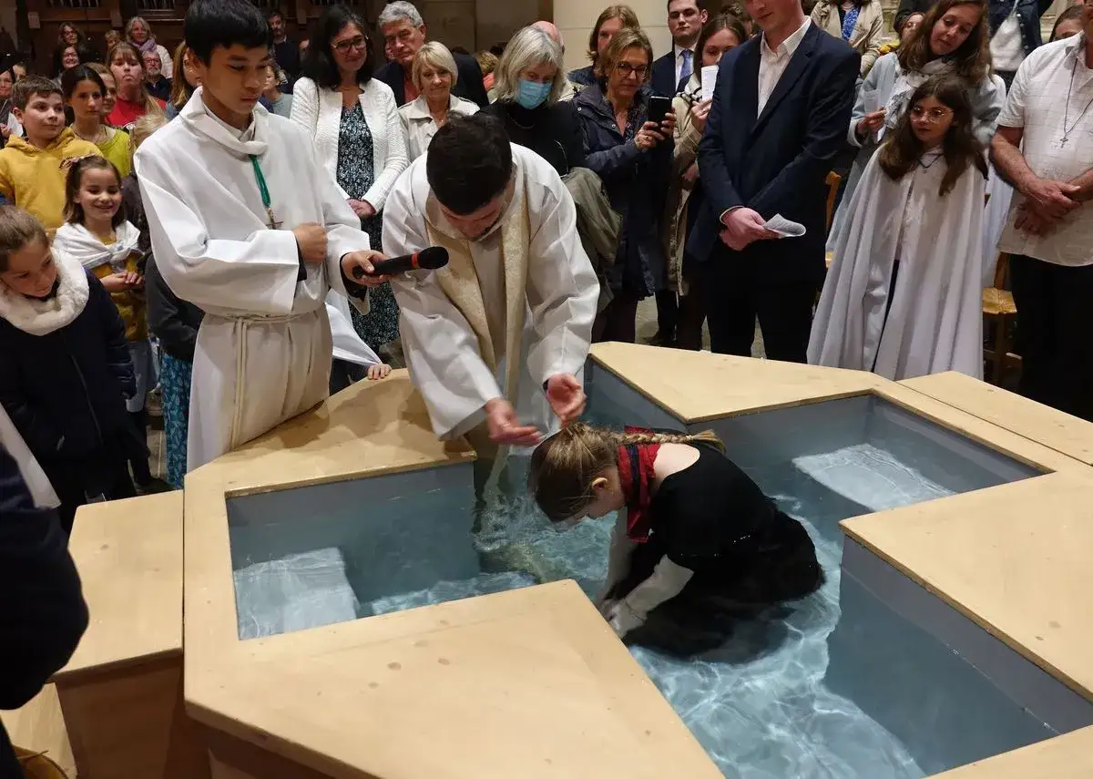 Cérémonie de baptême : un prêtre verse de l'eau sur la tête d'une jeune fille dans un grand bassin. On distingue les trois types de baptême par la présence de plusieurs enfants et adultes.