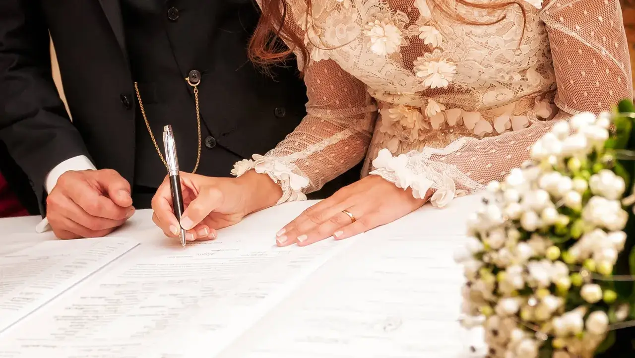 Un couple signe les registres lors de leur mariage civil à la mairie. La mariée porte une robe en dentelle, l'alliance est visible.