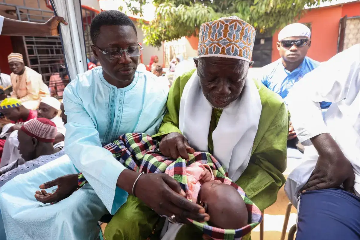 Un vieil homme et un jeune homme célèbrent un baptême au Sénégal, tenant un bébé enveloppé dans une couverture colorée.