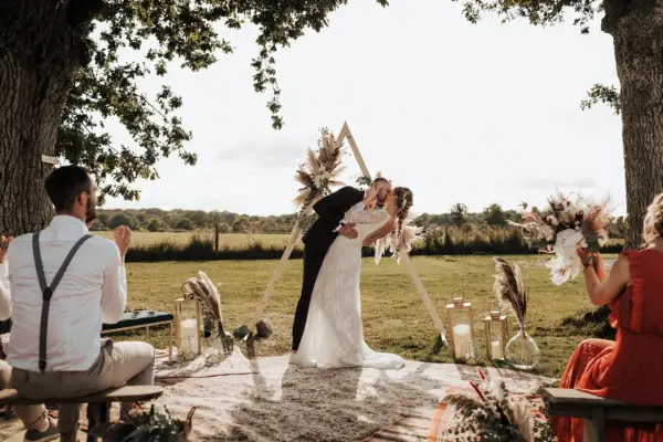 Un couple s'embrasse lors d'une **cérémonie laïque extérieur**. Un arche triangulaire décoré de fleurs et de plumes se dresse derrière eux.