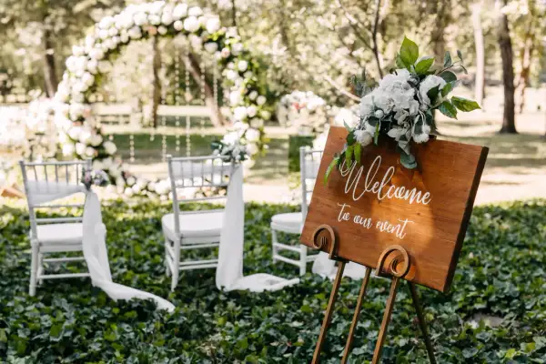 Bienvenue à notre mariage industriel en plein air. Arches florales, chaises blanches et panneau de bienvenue en bois.