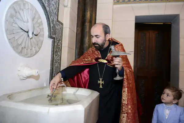 Un prêtre bénit l'eau pour un baptême arménien, un jeune garçon en costume bleu observe attentivement.