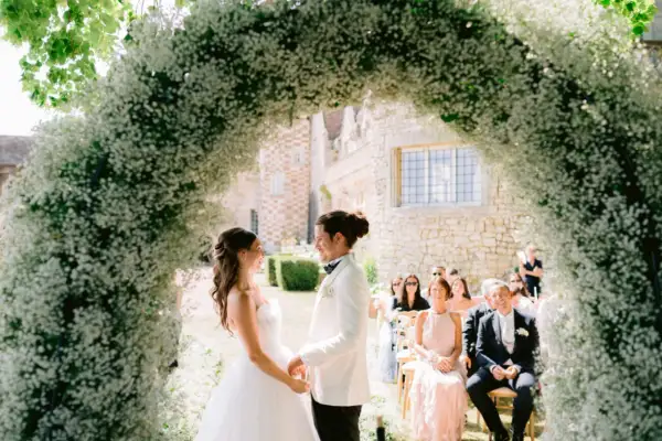 Un couple se tient sous une arche de fleurs blanches lors d'une cérémonie en plein air. L'homme, le parrain de cœur, porte un costume blanc.