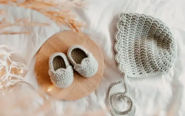 Doua nouveau-né en crochet gris, des chaussons sur un plateau en bois et un bonnet assorti, prêts pour une nouvelle aventure.