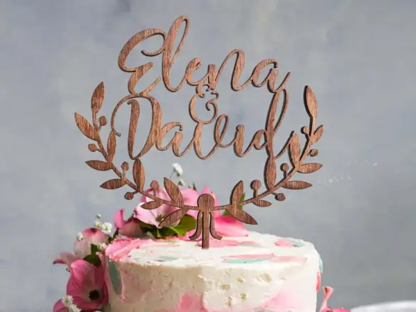 Topper gateau mariage en bois avec les prénoms "Elena & David" entourés de feuillage.
