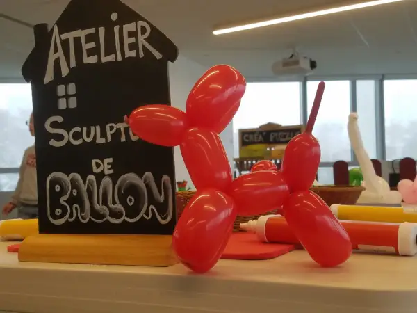 Un chien en sculpture ballon rouge, à côté d'un panneau "Atelier Sculpture de Ballon". Facile à réaliser !