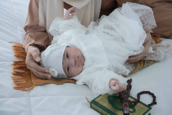 Un bébé en tenue blanche, entouré d'une mère, d'un tapis de prière, d'un Coran et de perles. Une scène évoquant les jours de naissance et leur sacrifice en Islam.