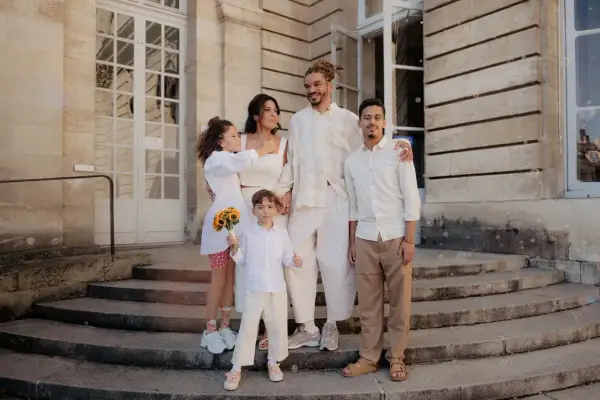 Famille réunie devant un bâtiment, le couple marié entouré de leurs enfants. La plus jeune porte un bouquet de tournesols, un vrai témoin mariage.