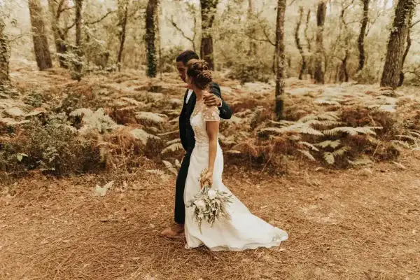Un couple se promène dans une forêt, la mariée porte une robe blanche et tient un bouquet. Un mariage DIY plein de charme.