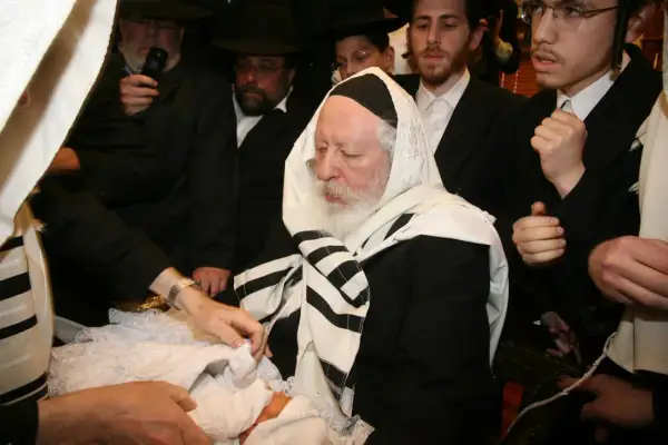 Cérémonie de circoncision juive. Un bébé est entouré d'hommes en habits traditionnels, dont un rabbin âgé.