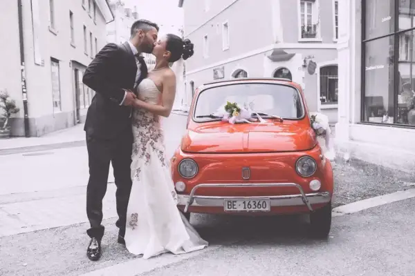Un couple s'embrasse devant une Fiat 500 rouge décorée, un doux souvenir de leur mariage à deux à l'étranger.